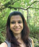 Sristi Kamal smiles against a green background of forest. She has brown hair with a red tinge. 