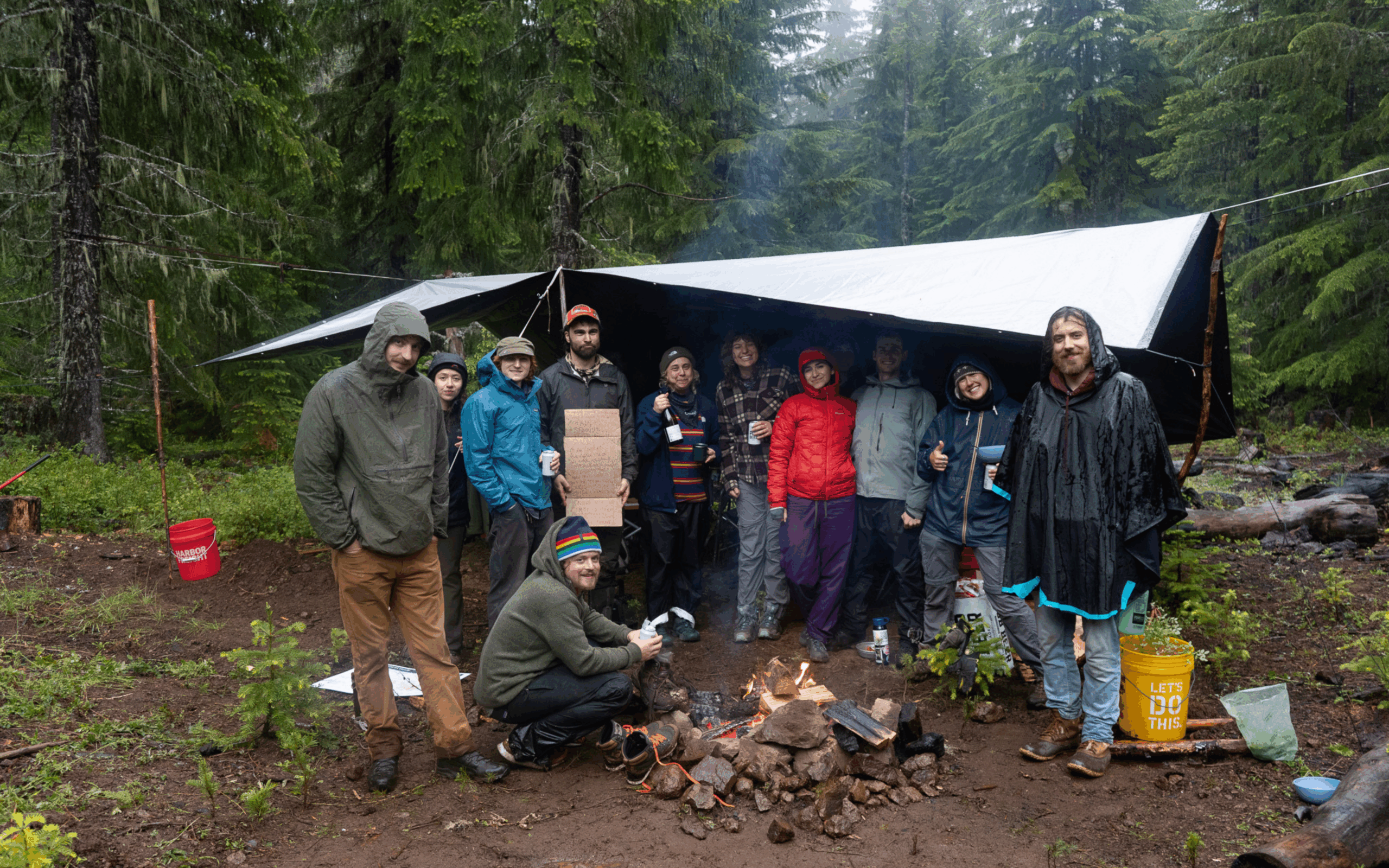 People in raincoats gather under a tarp and around a campfire on a rainy day.